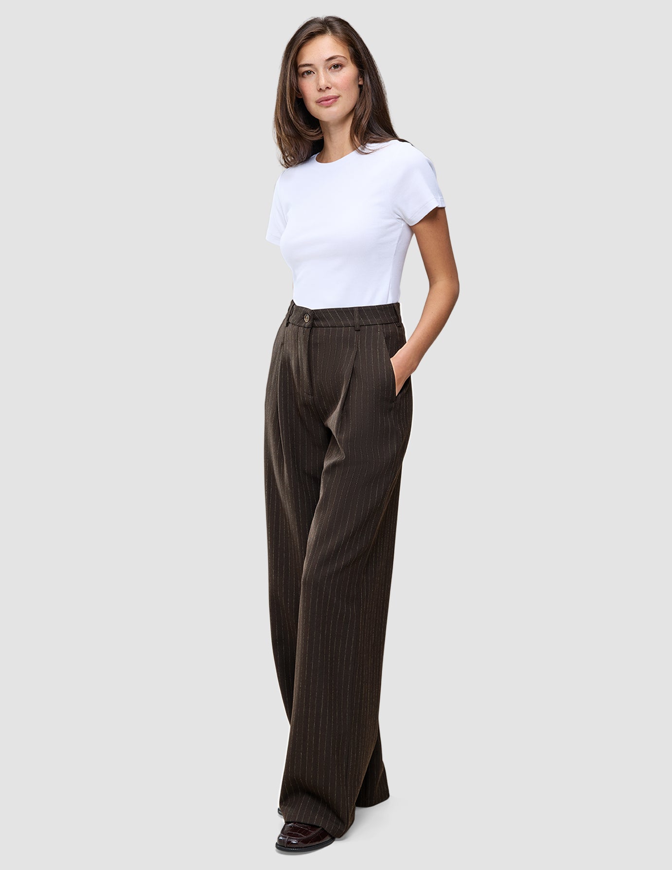 Eine Frau, die ein weißes T-Shirt und die Serene Pants Wide Walnut Herringbone trägt, die sich durch ein dezentes vertikales Streifenmuster und einen weiten Schnitt im skandinavischen Stil auszeichnen.